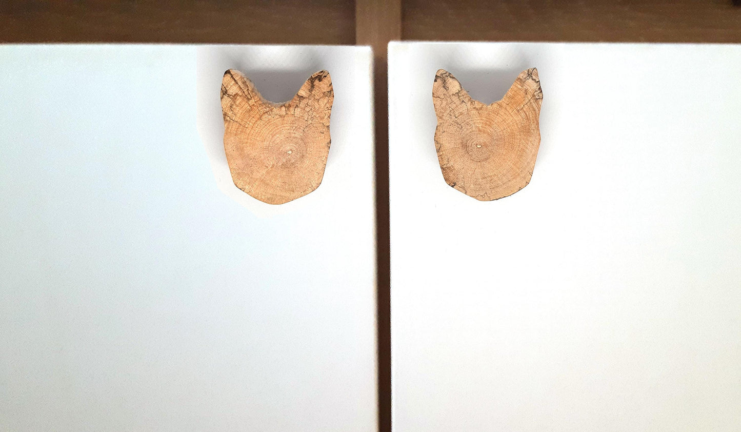 Paire de poignées en bois massif en forme de tête de chat, sculptées à partir de rondins de bois naturel avec leurs cernes visibles. Sur des portes blanches, elles apportent une touche artisanale et originale au mobilier. Fabrication artisanale