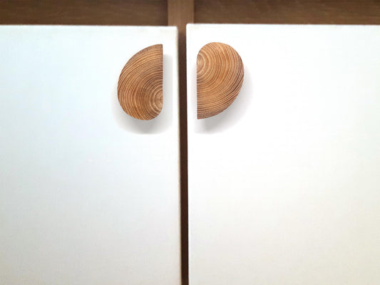 Vue rapprochée de deux poignées de placard en bois clair, en forme de demi-lune, montées sur des portes blanches. Leur position symétrique crée une esthétique circulaire harmonieuse une fois les portes fermées. Idéal pour une inspiration déco alliant simplicité, fonctionnalité et élégance naturelle. Parfait pour les recherches liées aux tendances de design d&#39;intérieur en bois et aux poignées de meuble contemporaines.