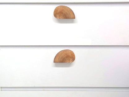 Détail de poignées de meuble en bois de forme semi-circulaire fixées sur deux panneaux blancs. L’alignement parfait de ces poignées crée un effet visuel circulaire esthétique, soulignant un style épuré et nordique. Cette solution de design unique est idéale pour des cuisines, dressings ou rangements modernes. Un exemple inspirant pour l’aménagement intérieur et la décoration avec des matériaux naturels.